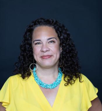 Ralina Joseph smiles in this headshot and wears a yellow flutter-sleeve blouse, a blue necklace, and long circle earrings. 