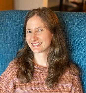 Adair Rounthwaite sits on a blue chair wearing a pink sweater. 