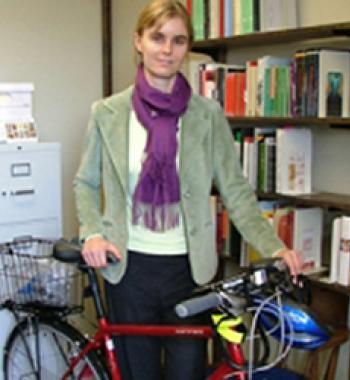Susan Gaylard stands behind a red bicycle and in front of a bookcase wearing a light-green jacket and purple sweater.