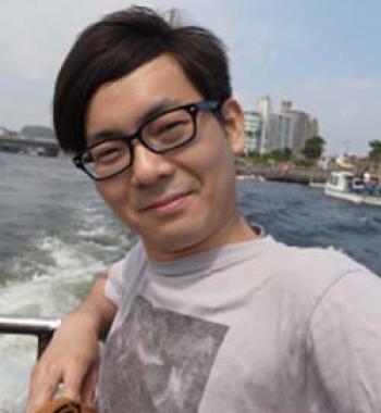 Yuta Kaminishi stands in front of a body of water wearing glasses and a grey shirt.