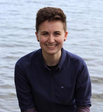 Profile picture of Sarah Brucia Breitenfeld: a person with short brown hair in a blue button up sitting in front of a lake.