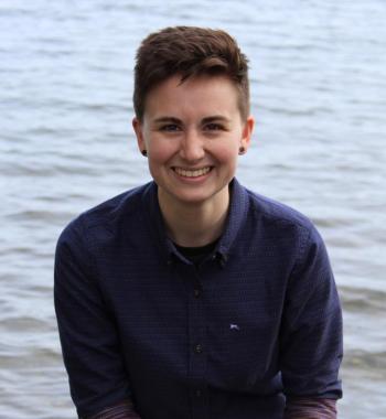 Profile picture of Sarah Brucia Breitenfeld: a person with short brown hair in a blue button up sitting in front of a lake.