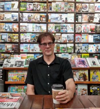 Image of Aaron Carpenter, male, sitting at a wooden table, holding a coffee cup. Magazines are displayed on shelves behind him.