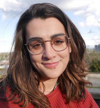 Close-up photo of a smiling woman with brown hair wearing round glasses and a red sweater. Part of the Seattle skyline is in view behind her.