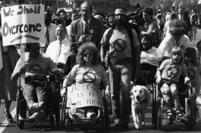 A black and white photo of a group of people walking and in wheelchairs protesting with signs.
