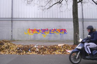 Yellow, blue, and red writing on a grey wall behind a gutter piled high with autumn leaves and street litter. On the right is a tall bare tree and an old Berliner biker zipping into the frame.