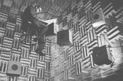 A scientist takes notes while sitting in a chair while insdie the Murray Hill Anechoic Chamber, which has hanging speakers and walls made of grids. 