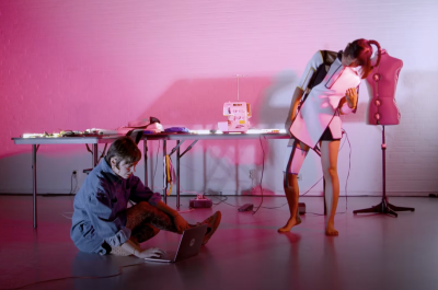 In a magenta lit room one woman sits on the ground with a laptop and the other stands while wearing a white futuristic garment.