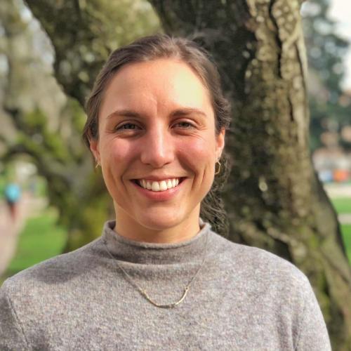 A portrait of Erika Versalovic standing in front of a tree wearing a grey sweater.