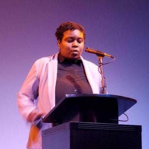Picture of Rasheena at Hugo House on stage reading writing in front of microphone on at podium.