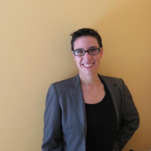 Picture of Sarah Levin-Richardson standing in front of a beige wall wearing glasses, a blazer, and a black shirt.