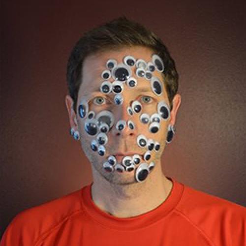 Ted Hiebert sits in front of a dark wall wearing an orange shirt and googly eyes all over his face.