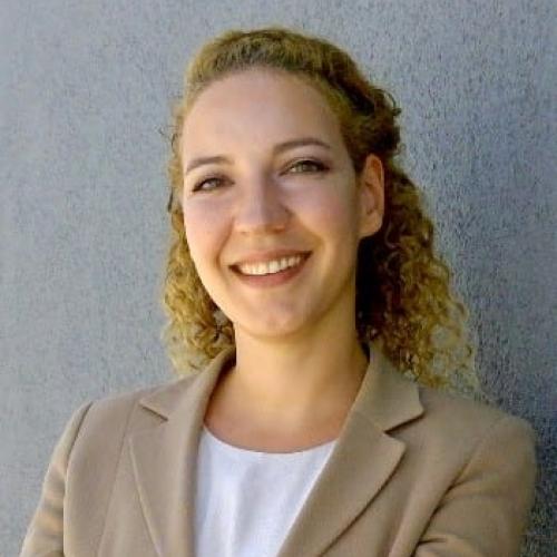 Ayda smiling at the camera in a beige blazer and white shirt, curly hair pulled halfway back and a gray background