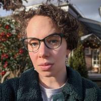 profile of Kaitlyn Boulding in glasses, close-up to the camera, wearing a jacket with a sky, trees, and buildings blurred in the background