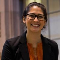 Profile of Katia smiling at the camera in a dark blazer and orange shirt