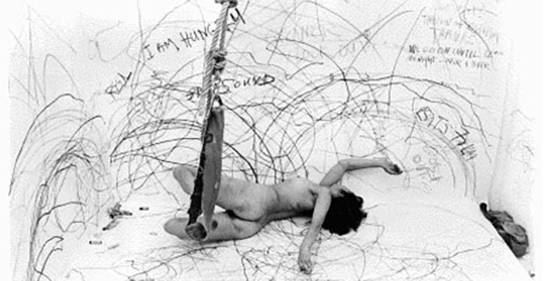 Photograph of Carolee Schneemann naked drawing on a room-sized paper and attached to a swing.