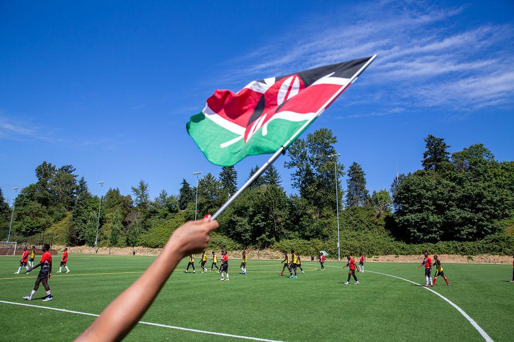 Hand holding a Kenyan flag with a scoccer field in the background