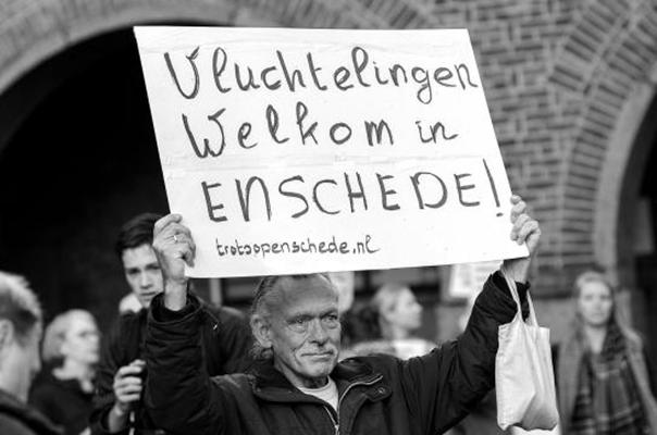 A black and white photo of a man holding a sign written in Dutch that reads Refugees Welcome in Enschede