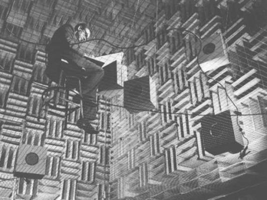 A scientist takes notes while sitting in a chair while insdie the Murray Hill Anechoic Chamber, which has hanging speakers and walls made of grids. 