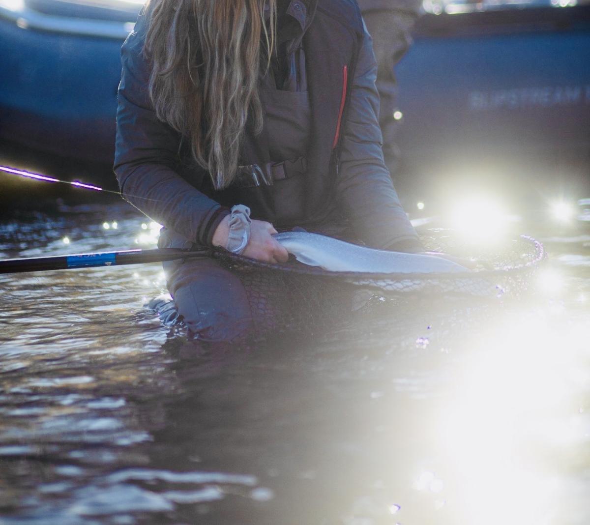 A figure sitting in water holds a fish inside of a net. The water is reflecting the sun