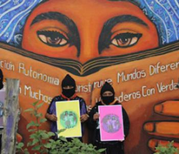 Three people wearing black hats and black masks hold signs while standing in front of a mural depicting a woman with long hair reading a book.