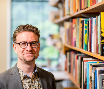 Christian Novetzke in glasses, a grey suit, and a print colored shirt. He is standing next to a line of books on shelves.