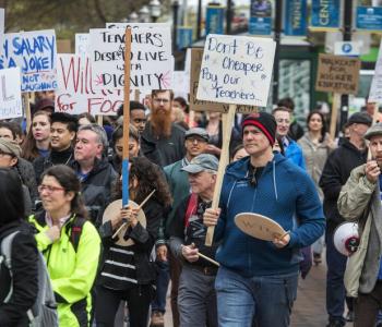 A group of people protest by walking together on a street, holding signs that protest salaries at Seattle-area community colleges.