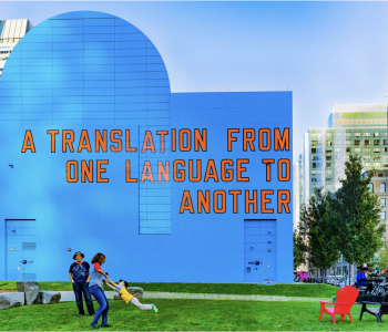 "A Translation From One Language to Another" is in orange text on a building painted bright blue. City skyscrapers are behind the building and people play in a grassy area in front of the building. The skies are sunny and blue.