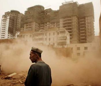Medium shot of a Uyghur man standing in Xinjiang, with dust and buildings in the background. Photo courtesy Carolyn Drake