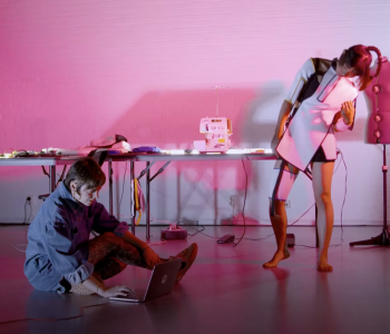 In a magenta lit room one woman sits on the ground with a laptop and the other stands while wearing a white futuristic garment.