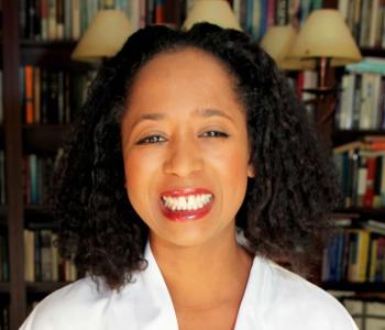 profile photograph of Jasmine Mahmoud: shoulder length curly hair, red lipstick, and a large smile with books in the background