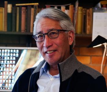 Profile photograph of Shawn Wong. He is Asian and male-presenting with greying short hair, black rimmed glasses, and a smile. He's wearing a shirt under a jacket with bookshelves in the background.
