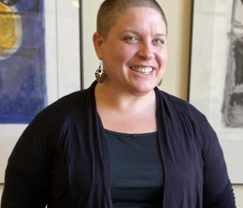 Julie stands in natural light smiling brightly. A thick white woman with a shaved head, she wears a black top and cardigan and large silver and black earrings. Behind her is a cream-colored wall with the framed edges of art pieces hung on each side of her.