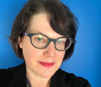 Portrait of Leigh Mercer wearing glasses and a dark jacket standing in front of a blue wall.