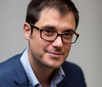 P. Joshua Griffin sits in front of a gray wall wearing glasses, a blue shirt, and a dark jacket.