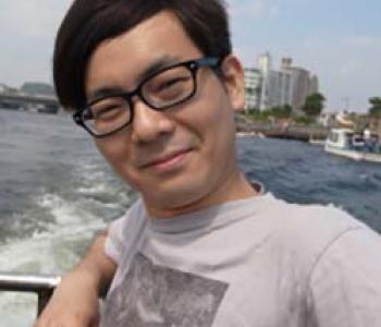 Yuta Kaminishi stands in front of a body of water wearing glasses and a grey shirt.