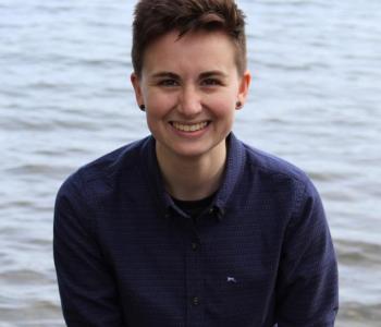 Profile picture of Sarah Brucia Breitenfeld: a person with short brown hair in a blue button up sitting in front of a lake.