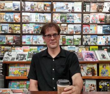 Image of Aaron Carpenter, male, sitting at a wooden table, holding a coffee cup. Magazines are displayed on shelves behind him.