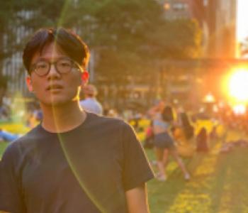 medium close-up of Yandong. He is on the left of the frame in a black t-shirt looking at the camera. To the right is a light flare form the setting sun, while the background shows buildings and a park.