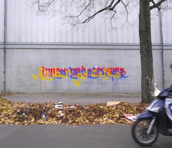 Yellow, blue, and red writing on a grey wall behind a gutter piled high with autumn leaves and street litter. On the right is a tall bare tree and an old Berliner biker zipping into the frame.