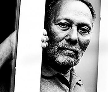 Black and white image of Stuart Hall looking at the camera from behind a pole.