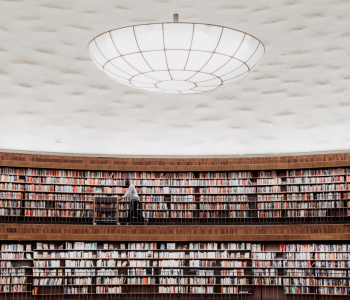 A large, circular library with two floors of bookshelves filled with colorful books. A person is pushing a cart along the upper floor walkway. The ceiling is dome-shaped and features a large, circular light fixture in the center. 