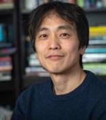 Jin-Kyu smiling at the camera in a blue sweater with blurred book stacks in the background