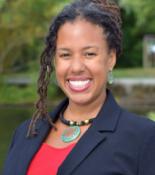 profile of Maya Smith smiling at the camera, hair pulled back with some curls along the left side, wearing a red top under a blue blazer with a necklace