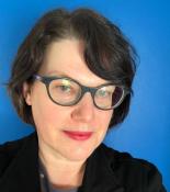 Portrait of Leigh Mercer wearing glasses and a dark jacket standing in front of a blue wall.