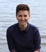 Profile picture of Sarah Brucia Breitenfeld: a person with short brown hair in a blue button up sitting in front of a lake.